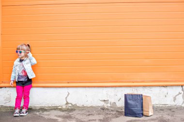 Little stylish girl in sunglasses with craft bags on shopping trip on an orange background. Shopaholic dances against the wall of the commercial center. Girl happy shopping on Black Friday. Copy space
