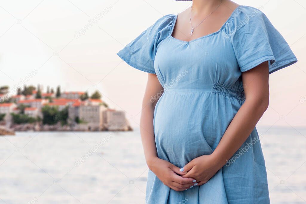 A pregnant woman traveler hugs her baby in her stomach near the island of Sveti Stefan in Montenegro. Close-up view Girl in a blue denim, fashionable dress. Tourism and travel in pregnancy. Copy space