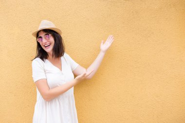Hasır şapkalı, yuvarlak güneş gözlüklü orta yaşlı bir kadın, gülüyor, beton turuncu duvarı işaret ediyor. Turist gevşek bir bej elbiseyle neşeli bir şekilde seyahat eder. Boşluğu kopyala, metin için yer aç. Mutlu kadın.