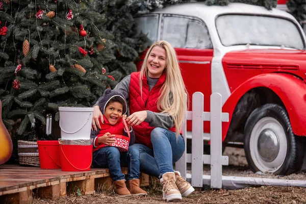 Caucasian mother hugs, kisses African American son, sitting by the Christmas tree, behind a white fence, a red car. Young woman strokes the head of boy who opens gifts from Santa Claus. Happy holidays