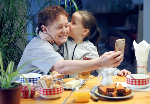 Grandma Granddaughter Sitting Kitchen Looking Photos Phones Taking Selfies Warm — Stockfoto