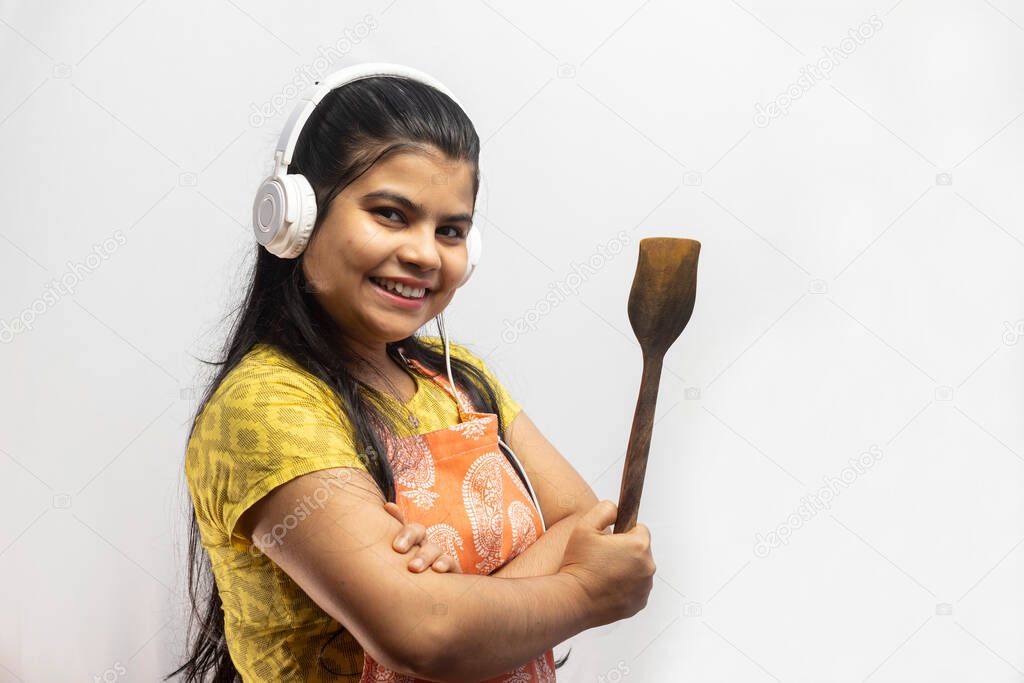 A pretty Indian housewife woman wearing cooking apron and headphone with wooden spatula in hand smiles on white background