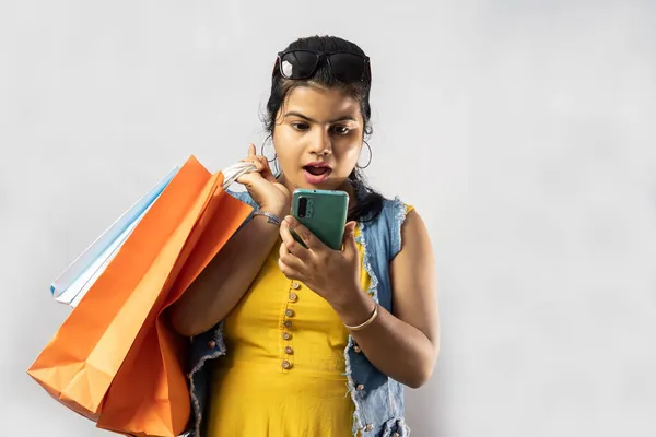 美しいですインド人女性で黄色のドレスでショッピングバッグ見ます彼女のスマートフォン上の白い背景 — ストック写真