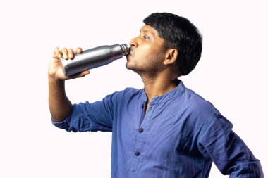 Mavi elbiseli genç Hintli bir adamın yüzü beyaz arka plandaki paslanmaz çelik şişeden su içiyor.