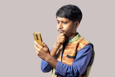 Mavi elbiseli genç bir Hintli. Düşünceli bir yüzü var. Gri arka planda akıllı bir telefon tutuyor.