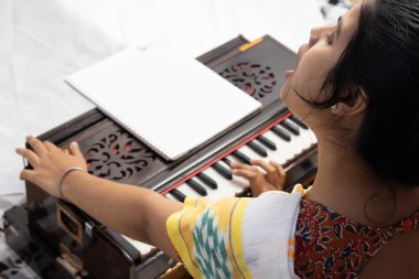 Sari giymiş bir Hintli kadın beyaz arka planda şarkı söylüyor ve harmonyum çalıyor.