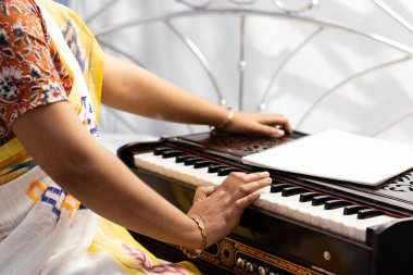 Sari giymiş bir Hintli kadın beyaz arka planda şarkı söylüyor ve harmonyum çalıyor.