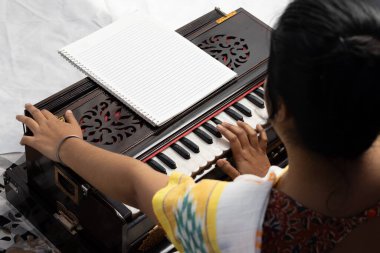 Sari giymiş bir Hintli kadın beyaz arka planda şarkı söylüyor ve harmonyum çalıyor.