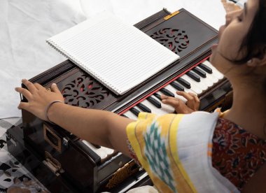 Sari giymiş bir Hintli kadın beyaz arka planda şarkı söylüyor ve harmonyum çalıyor.