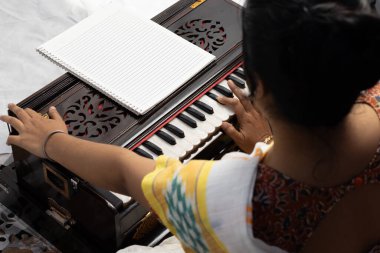 Sari giymiş bir Hintli kadın beyaz arka planda şarkı söylüyor ve harmonyum çalıyor.