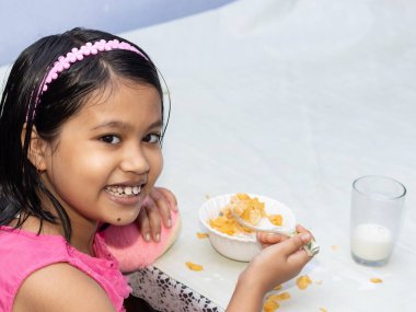 Hintli bir kız çocuğu gülümseyen yüzüyle mısır gevreği ve süt yiyor ve kameraya bakıyor.