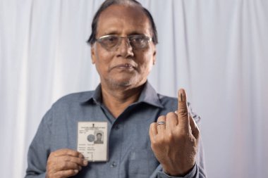 Beyaz arka planda elinde seçmen kartıyla gülümseyen Hintli yaşlı bir adam.