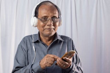 Beyaz arka planda kulaklık takan yaşlı bir Hintli akıllı telefon kullanıyor.