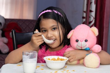 Hintli bir kız çocuğu sütle mısır gevreği yiyor ve gülümseyen yüzüyle kameraya bakıyor.