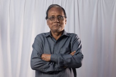 Yaşlı bir Hintli beyaz arka planda kolları kapalı kameraya bakıp gülümsüyor.