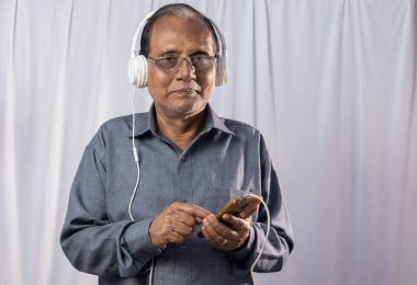 Beyaz arka planda kulaklık takan yaşlı bir Hintli akıllı telefon kullanıyor.