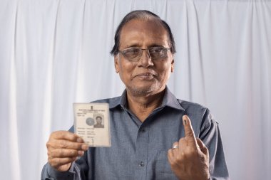 Beyaz arka planda elinde seçmen kartıyla gülümseyen Hintli yaşlı bir adam.