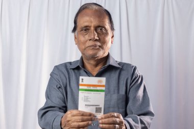 Beyaz arka planda elinde Aadhaar kartıyla gülümseyen Hintli yaşlı bir adam.