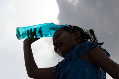 Serinlemek için gökyüzünün altındaki mavi plastik şişeden su sıçratan bir kızın silüeti.