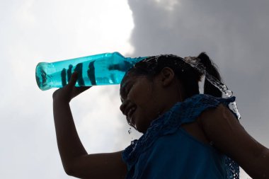 Serinlemek için gökyüzünün altındaki mavi plastik şişeden su sıçratan bir kızın silüeti.