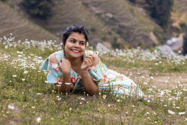 A cheerful smiling Indian girl lying on field full of calendula flowers looks away with a flower in hand