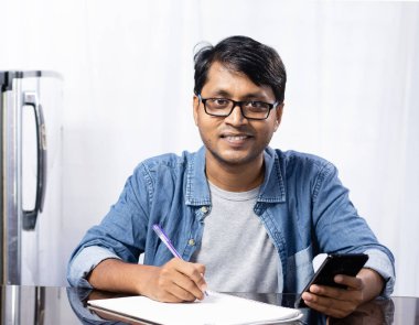 Beyaz arka planda akıllı bir telefonla evde çalışırken gülümseyen yüzüyle kameraya bakan Hintli genç bir erkek.