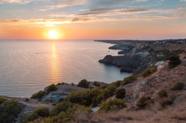 Gün batımında Cape Fiolent 'te panoramik manzara. Ufukta büyük, kırmızı bir güneş bulutlu, pembe bir gün batımına karşı batıyor. Alacakaranlık güneşi kayalık uçurumlarda parlıyor. Kayalıkların altındaki Karadeniz.