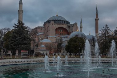 Hagia Sophia and the large round fountain are located on Sultanahmet Square. Water jets are a little blurry. The temple has high minarets and several large domes. The cathedral is made of red stone.