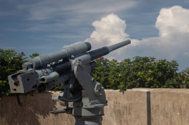 An old battered model of coastal gun of medium caliber with peeling paint. The weapon decorate shore as a sign of passed world war 2. This cannon used to defend fortifications against enemy warships.