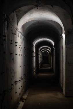 A long dark dimly tunnel of an abandoned military bunker or bomb shelter with gray concrete walls and metal parts sticking out them. The arched ceiling is illuminated by dusky lamps.