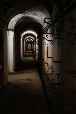 A long dark dimly tunnel of an abandoned military bunker or bomb shelter with gray concrete walls and metal parts sticking out them. The arched ceiling is illuminated by dusky lamps.