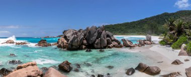 A high-resolution panorama of a tropical beach with huge stone formations in the middle. Turquoise water washes over the white sand beach. A rainforest of palm trees grows along the shore.