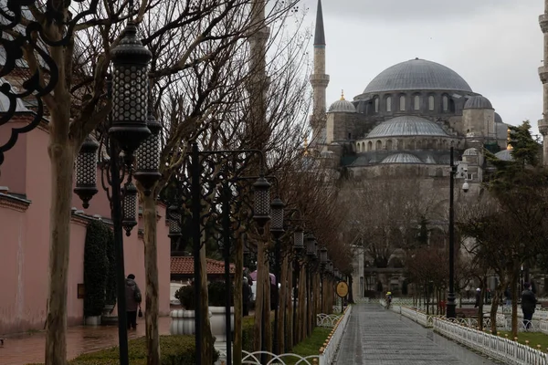 The Blue Mosque stands at the far end of a pavement walking alley with decorative lampposts. The lovely place is in Istanbul at the Sultan Ahmed square. It is made of gray stone, has high minarets.