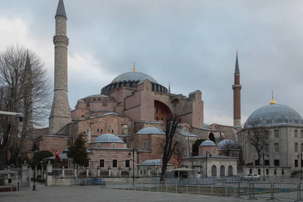 Hagia Sophia and the large round fountain are located on Sultanahmet Square. Water jets are a little blurry. The temple has high minarets and several large domes. The cathedral is made of red stone.
