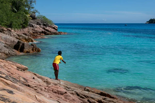 A young Seychelles fisherman is fishing on his home island. A boy kid is throwing fishing rod into crystal blue water of the ocean from rocky steep shore. This is the islanders' way of life