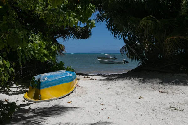 Upside down wooden coloured boat is on white sand against a backdrop of green tropical bushes on the beach. In the distance, through the arch of trees is seen blue ocean water and horizon line.