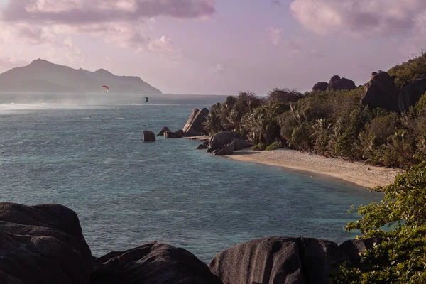 Panoramic view of a evening bay of Indian Ocean in the Seychelles with a couple of kite surfers. Huge granite stones on the beach. Shore is covered with huge round rocks and dense dusky jungles.  
