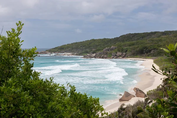 View from high viewpoint of the snow-white Grand Anse Beach on La Digue island in the Seychelles. Raging ocean washes over the wide shore. In the distance, huge granite cliffs covered with rainforest 