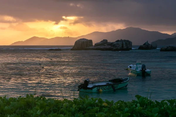 A seascape during sunset. Two small moored boats stand in turquoise water against an orange sunset sky. In the distance you can see rocks and the silhouette of an island.