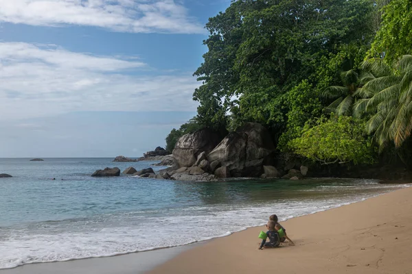 A couple of children, a boy and a girl, sit on the beach in the wet sand close to the sea. The kids are surrounded by lush tropical vegetation. The bay is fenced off by massive stones 