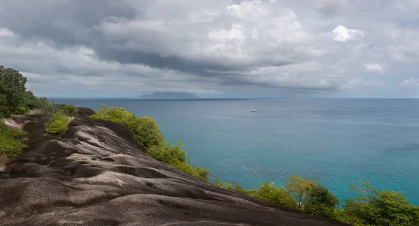 View from a high flat granite cliff overlooking the turquoise bay of the Indian Ocean in the Seychelles. The coastline is covered with huge round rocks, steep cliffs and trees