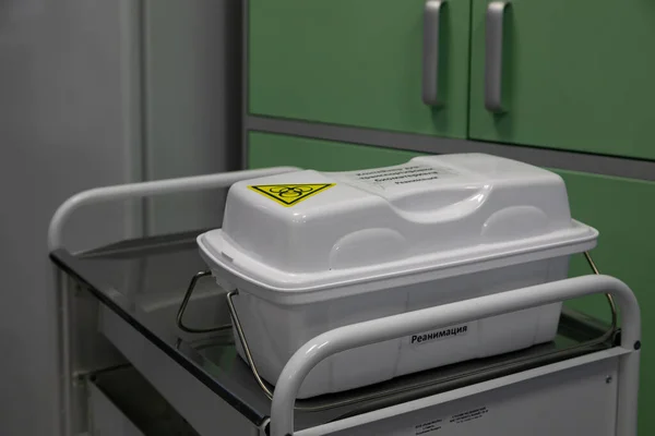 Close-up of a plastic container for collecting biological materials such as blood, urine and sputum. The container is grey and stands on a metal medical table in the background of a hospital setting.