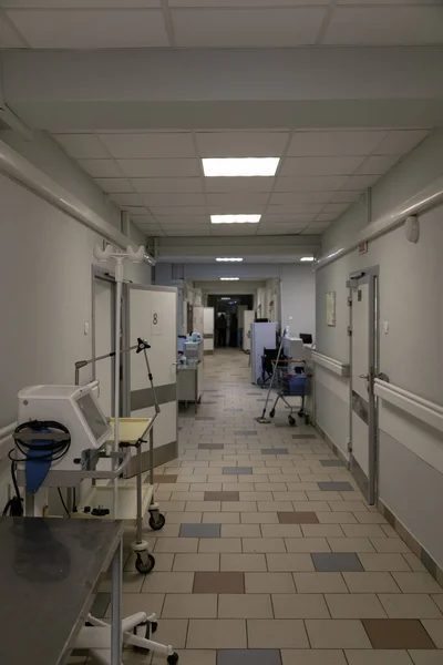 The hospital corridor of a Russian hospital. The doors to the wards are open, with a nurse's station between them. Medical equipment along the grey walls