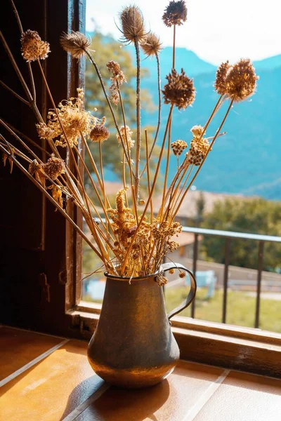 Metal vase with dried flowers near the window at dawn. Decoration of a house in the countryside