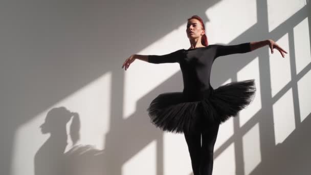 Балерина в черной туту изящно танцует против белой стены в ярком солнечном свете — стоковое видео