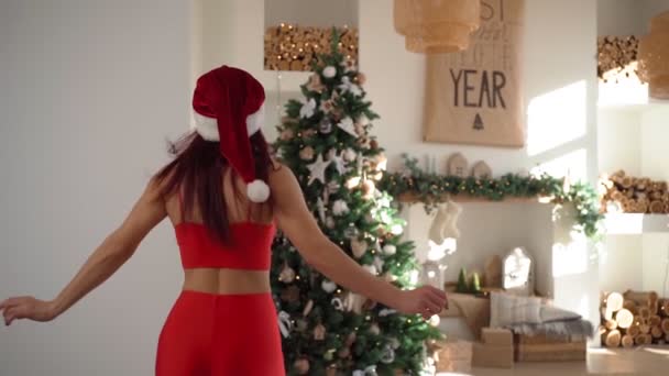 Mujer feliz en el sombrero de Papá Noel ejecuta el árbol de Navidad en el espacioso apartamento brillante — Vídeos de Stock