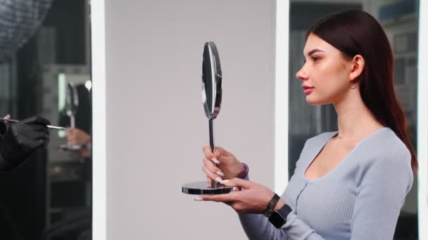 Vacker kvinna med perfekt hud ansikte ser sig själv i spegeln i skönhetssalong — Stockvideo
