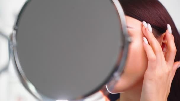 Mooie vrouw met perfecte huid gezicht ziet zichzelf in spiegel in schoonheid salon — Stockvideo
