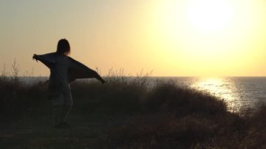 Genç bir kadın gün doğumunda meditasyon yapar, dans eder ve döner. Deniz kıyısında gün batımında. Bir kadının silueti, meditasyon, rahatlama, tatil, seyahat, deniz manzarası. Yoga dersleri, güneş ışınları. 4K video.