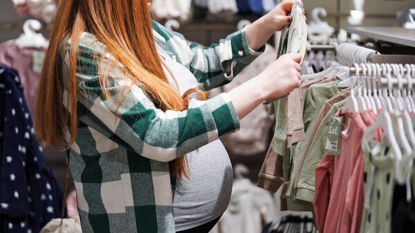 Крупный план живота беременной рыжеволосой женщины в магазине детских вещей, торговом центре, сезоне скидок и продаж. Будущая мать выбирает одежду для своего ребенка, подготовку к рождению ребенка